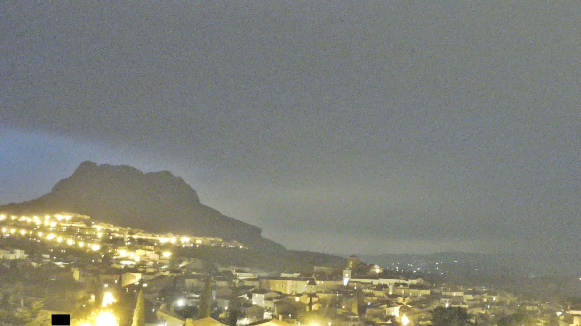 A nighttime view of a town nestled at the base of a dark mountain under a hazy, overcast sky.