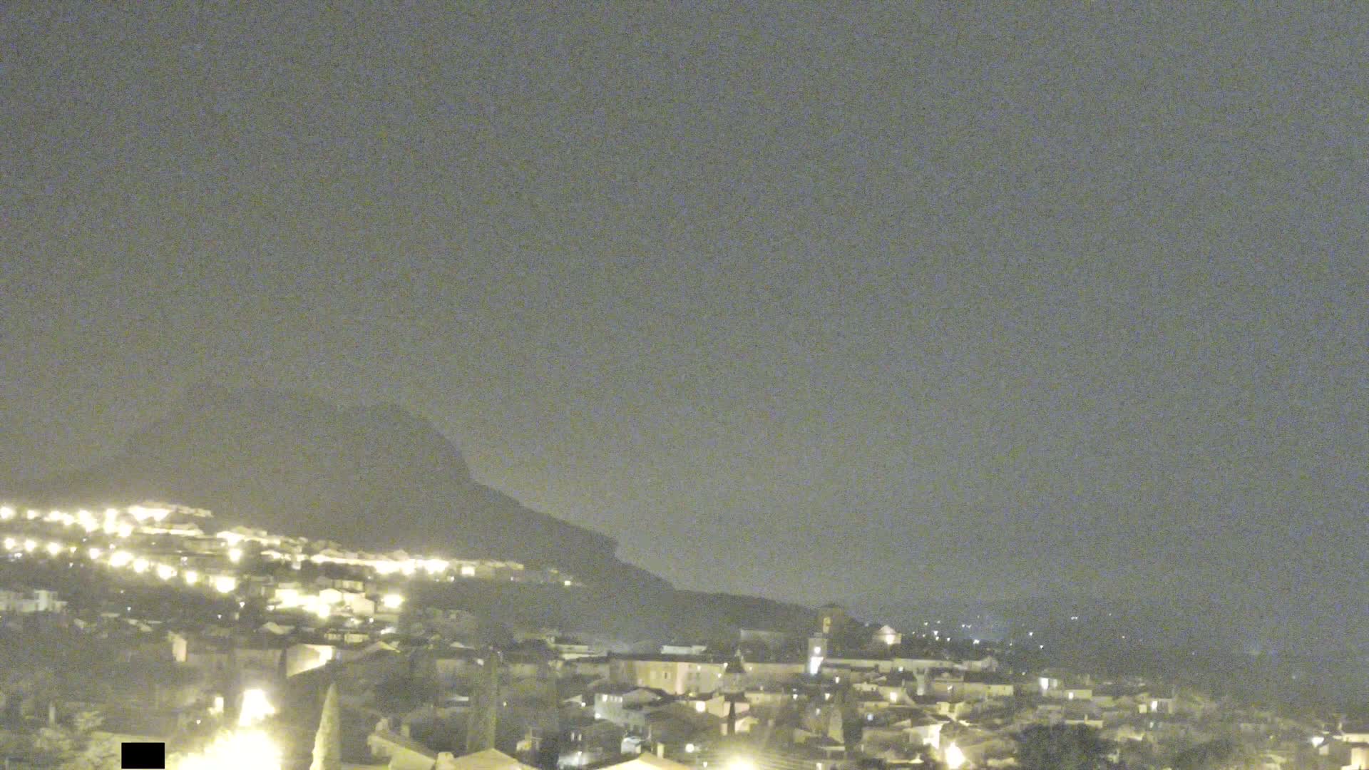 A nighttime view of a town nestled against a hill under a hazy sky.