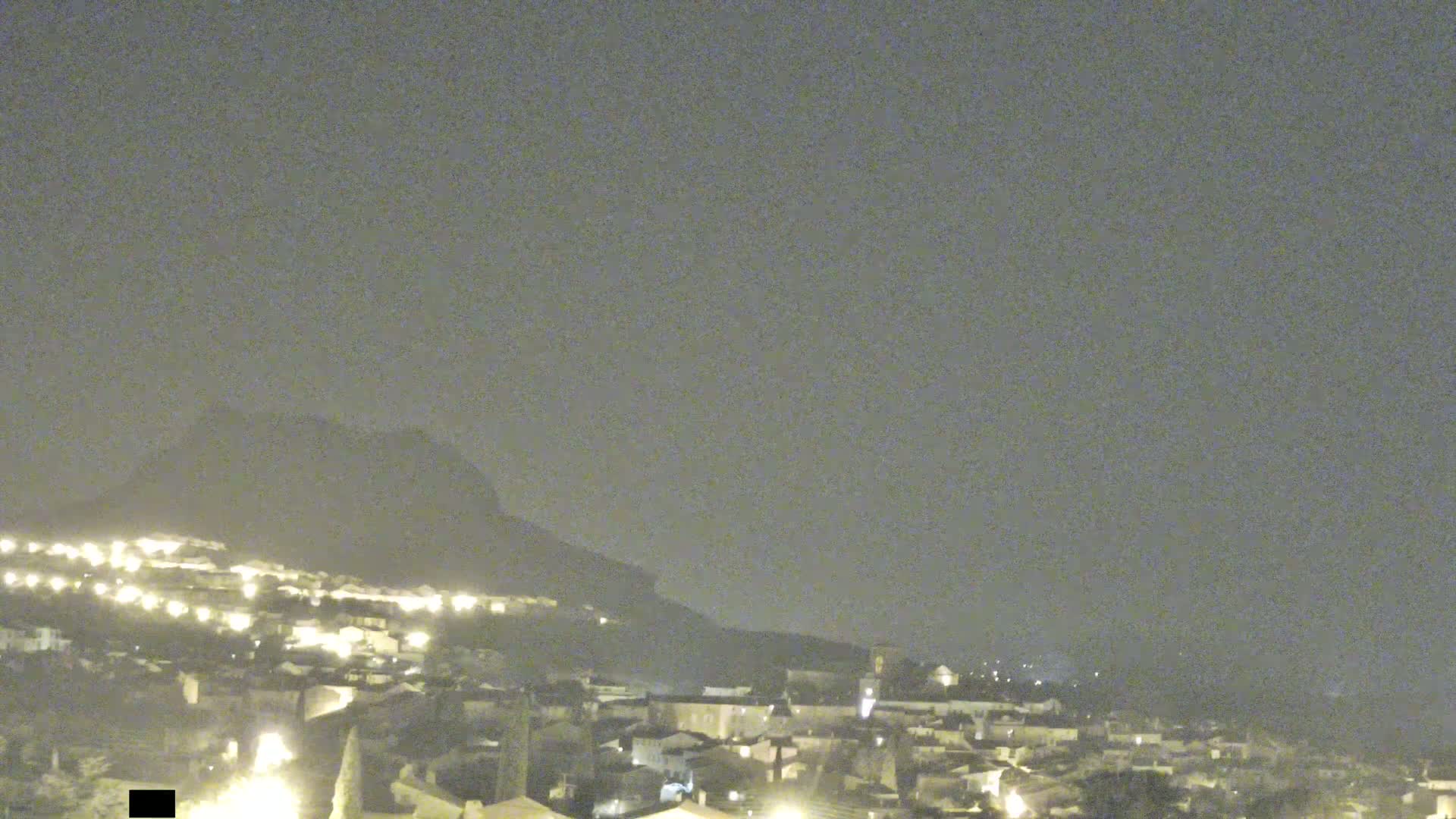 A nighttime view of a town nestled against a hill under a hazy sky.