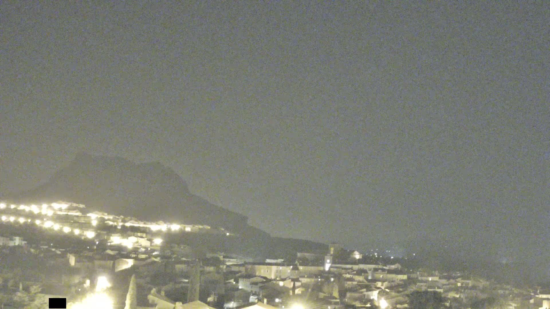 A nighttime view of a town nestled against a hill under a hazy sky.