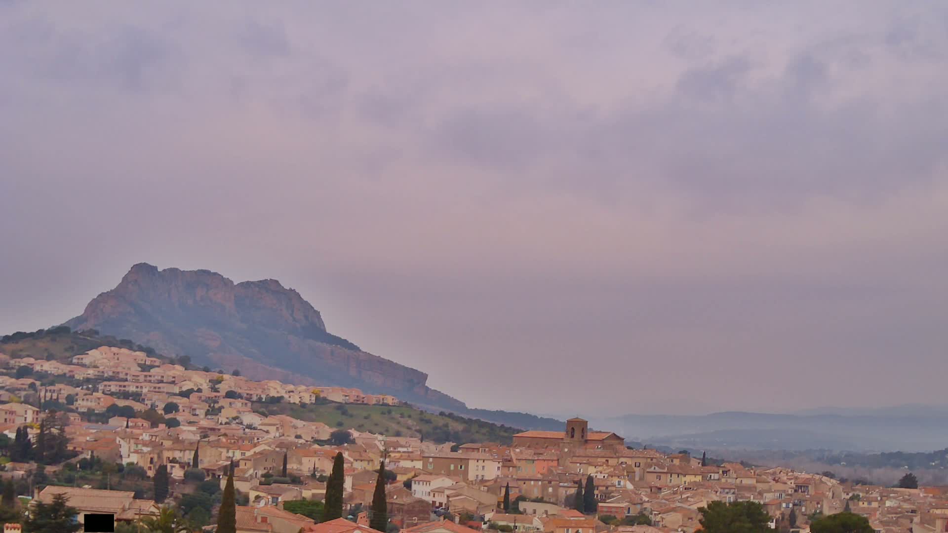 A hillside town of tan-colored buildings nestles beneath a large, dark mountain under a hazy, pale purple sky.