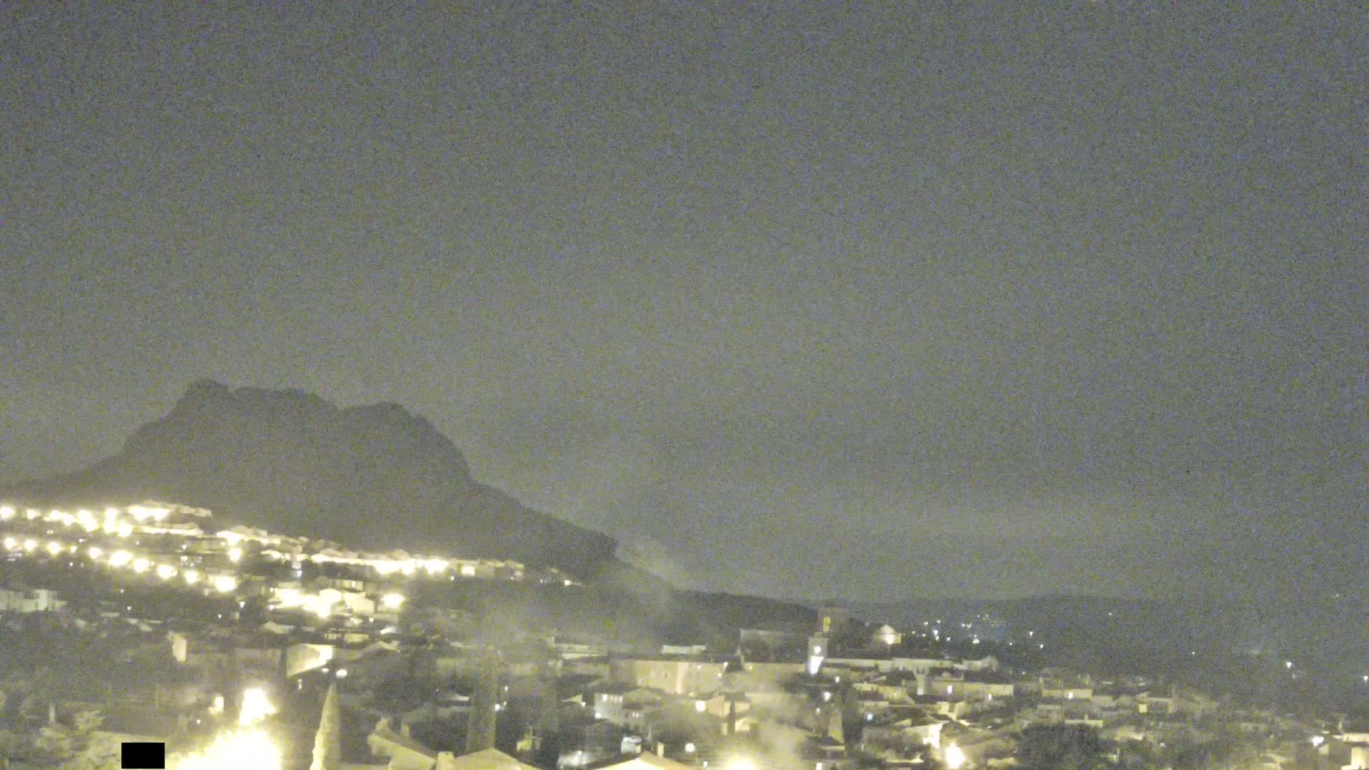 A nighttime view of a town nestled below a dark mountain under a cloudy sky.