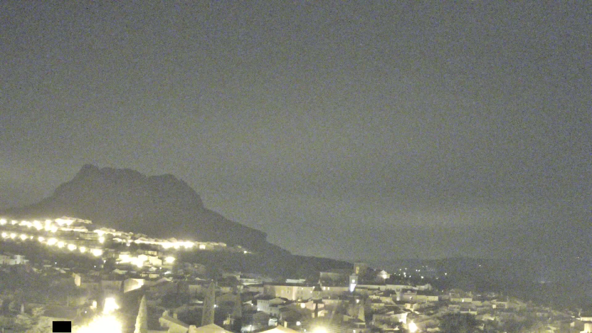 A nighttime view of a town nestled at the base of a dark mountain under a hazy sky.