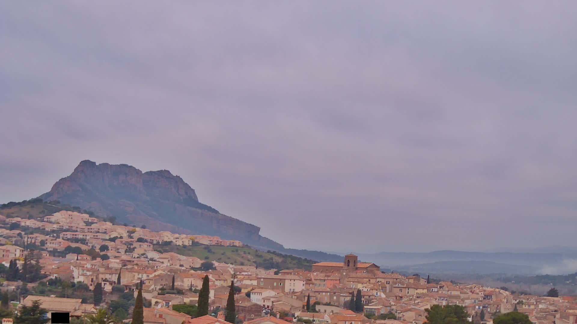 A hilltop town of light brown buildings nestles against a large, dark mountain under a pale, cloudy sky.