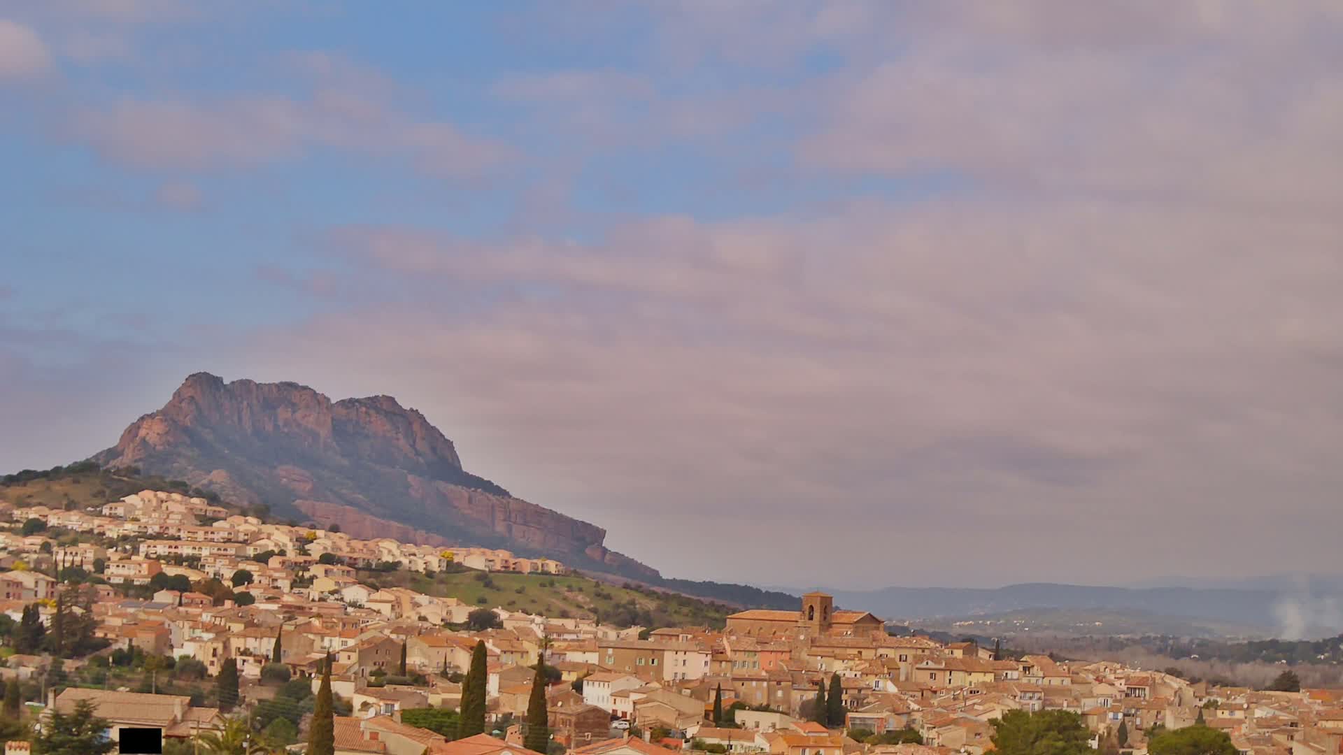 A sprawling town of tan-colored buildings nestles against a large, dark-brown mountain under a pale, cloudy sky.