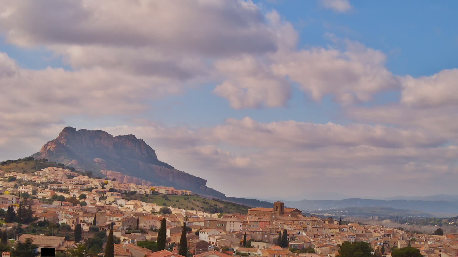 A town of tan buildings nestled on a hillside beneath a large, dark mountain under a partly cloudy sky.