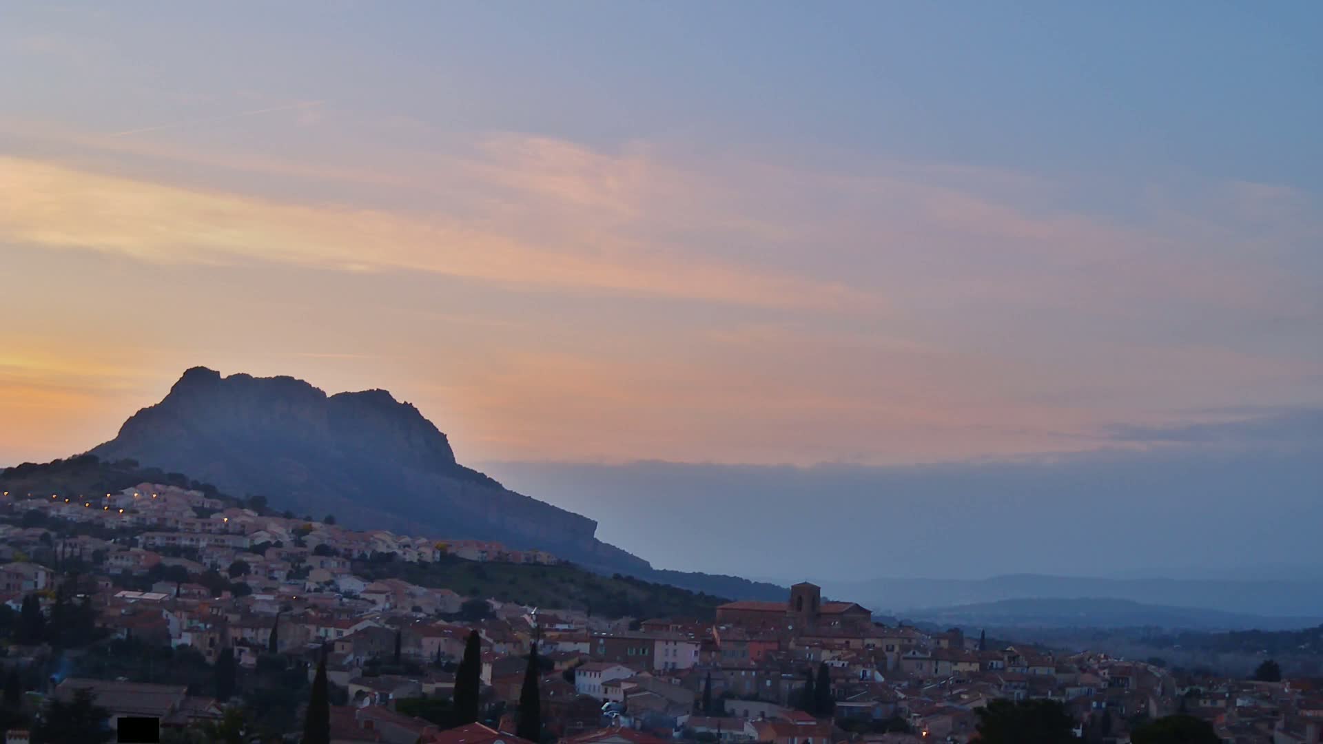 A village nestled at the base of a large, dark mountain under a soft, pastel sunset sky.