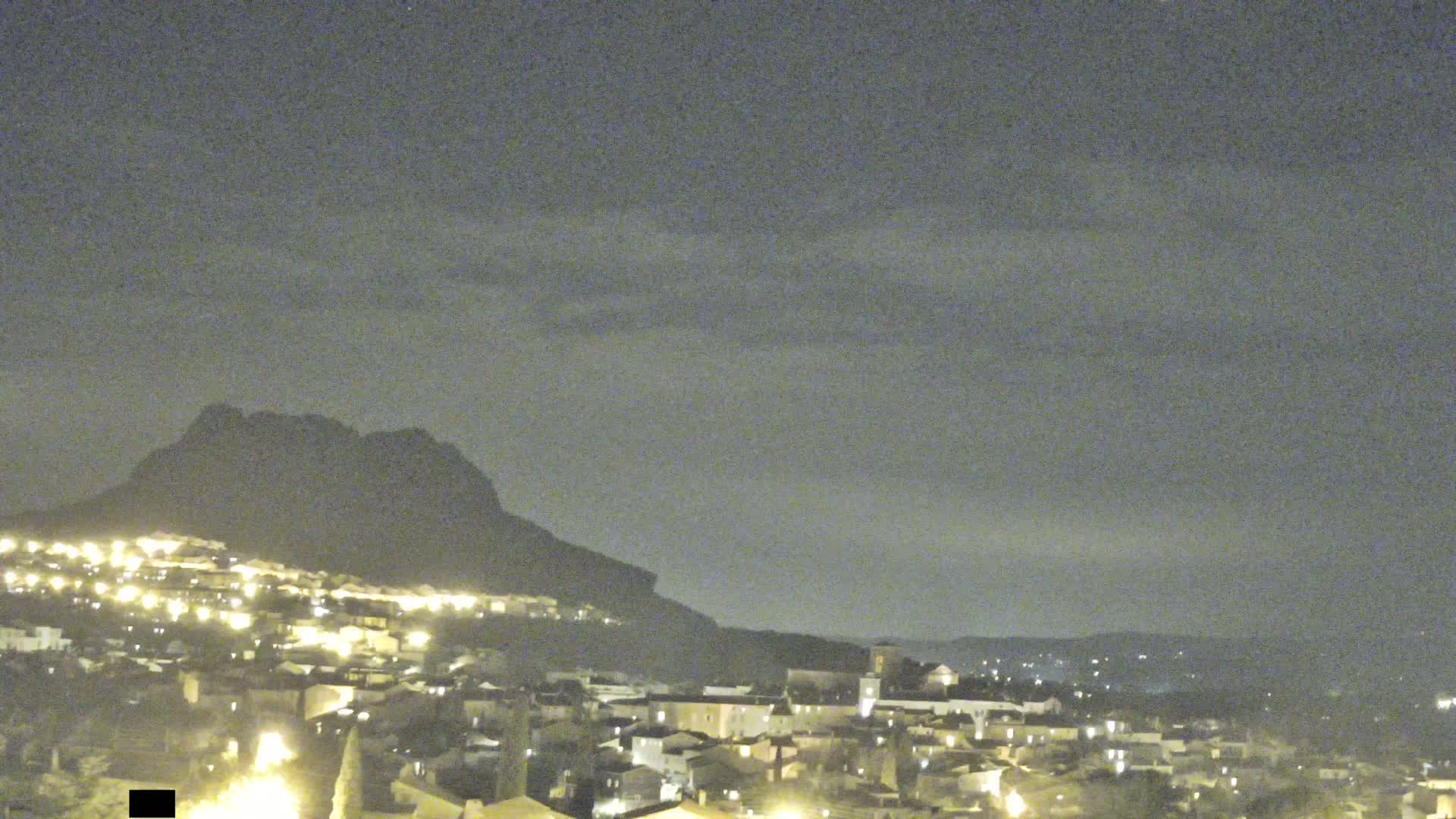 A nighttime view of a town nestled against a dark mountain under a mostly cloudy sky.