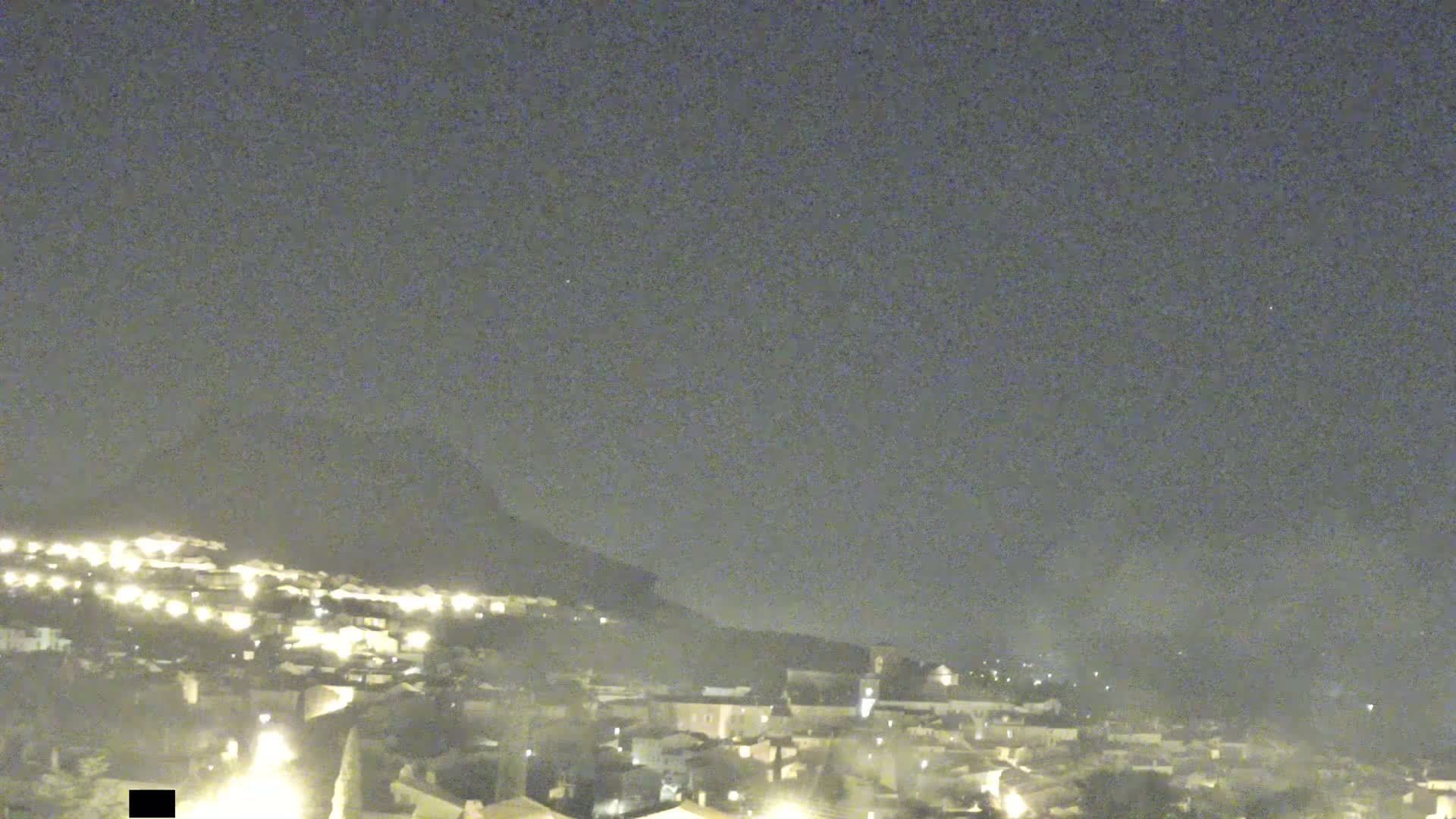 A nighttime view shows a small town nestled against a hill under a dark, overcast sky.