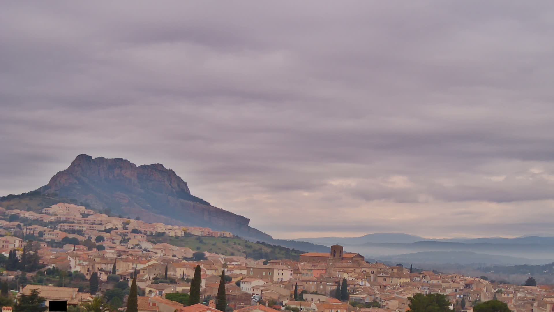 A sprawling town of tan and brown buildings nestles at the base of a large, dark mountain under a cloudy sky.