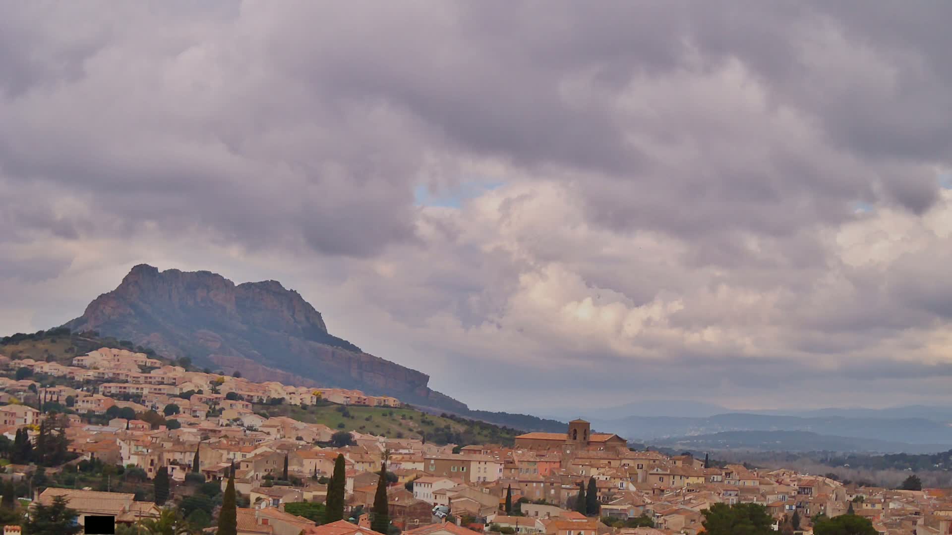 A hillside town with tan and brown buildings nestles beneath a large, dark mountain under a cloudy sky.