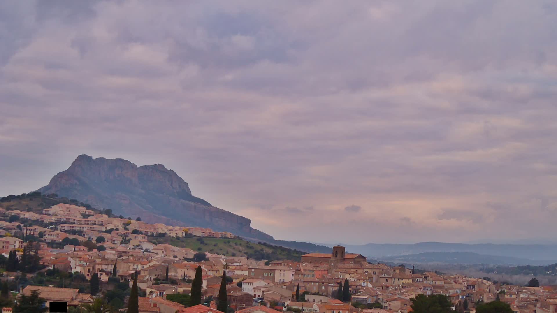 A hilltop town with tan and brown buildings nestles against a large, dark mountain under a cloudy, pale purple sky.