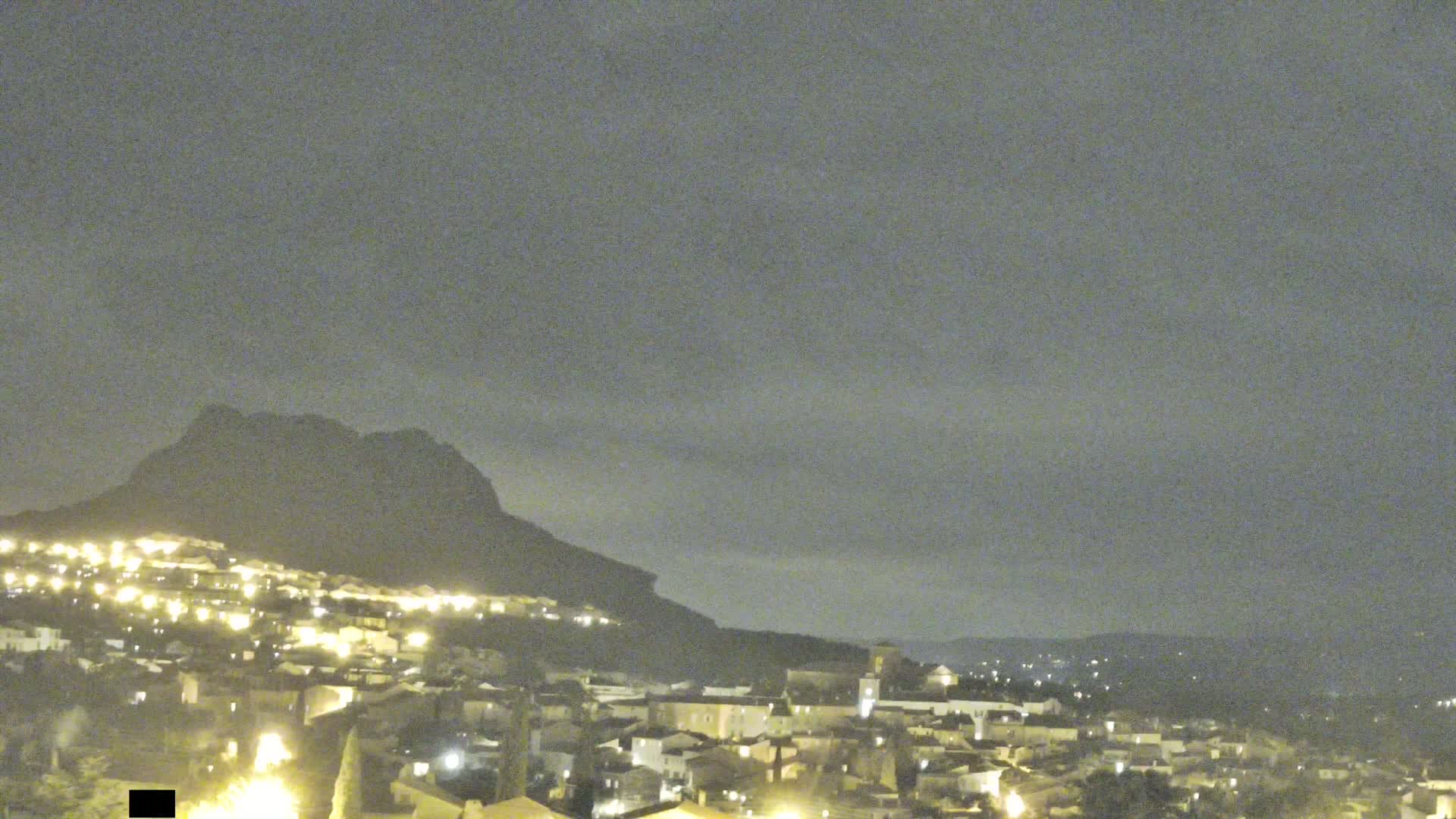 A nighttime view of a town nestled against a large, dark mountain under a cloudy sky.