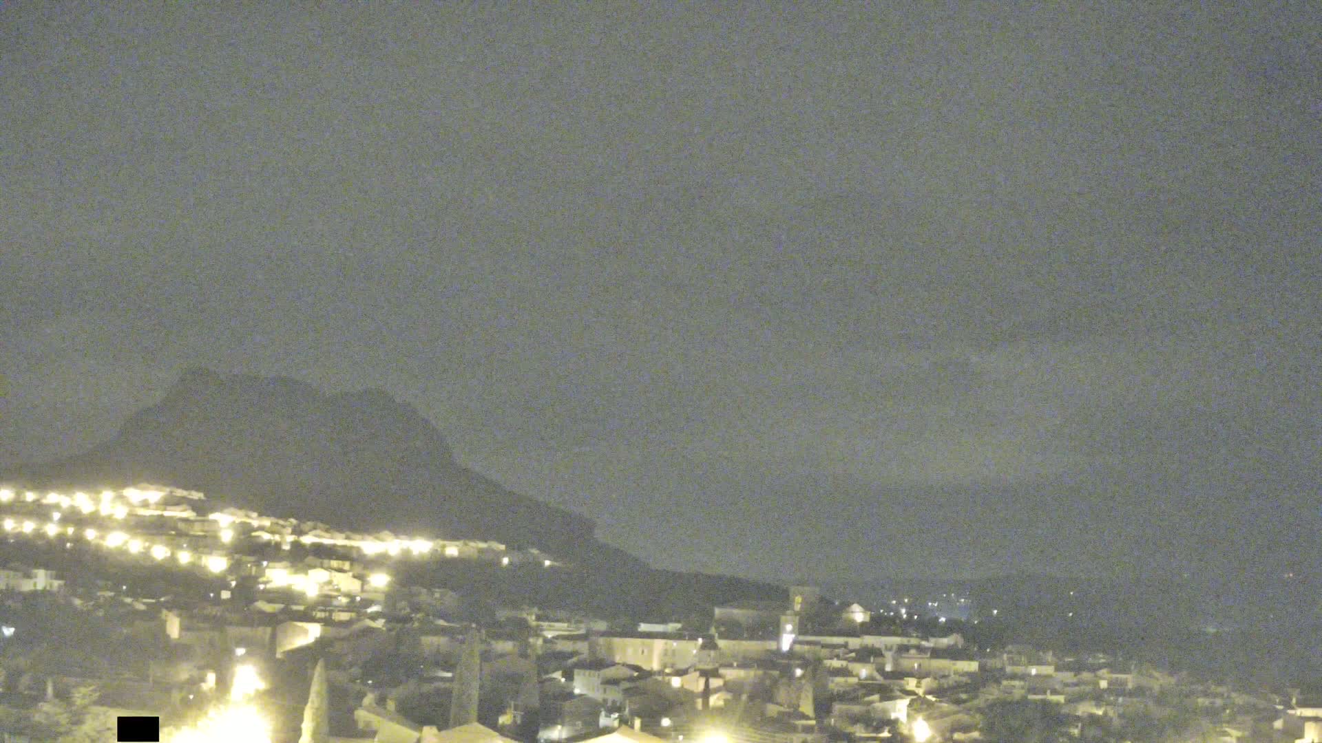 A nighttime view shows a dimly lit town nestled at the base of a dark mountain under a cloudy sky.