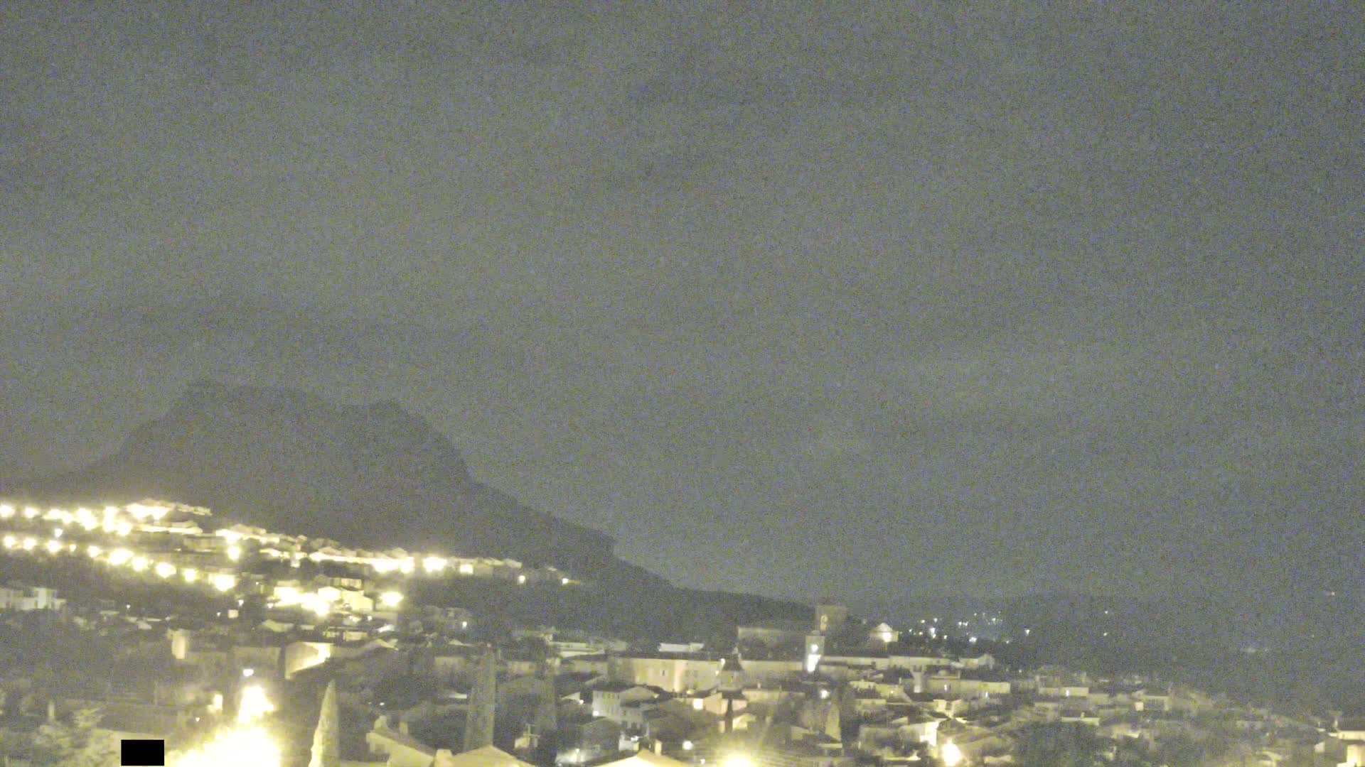 A nighttime view of a hilltop town illuminated by scattered lights, with a large, dark mountain in the background under a cloudy sky.