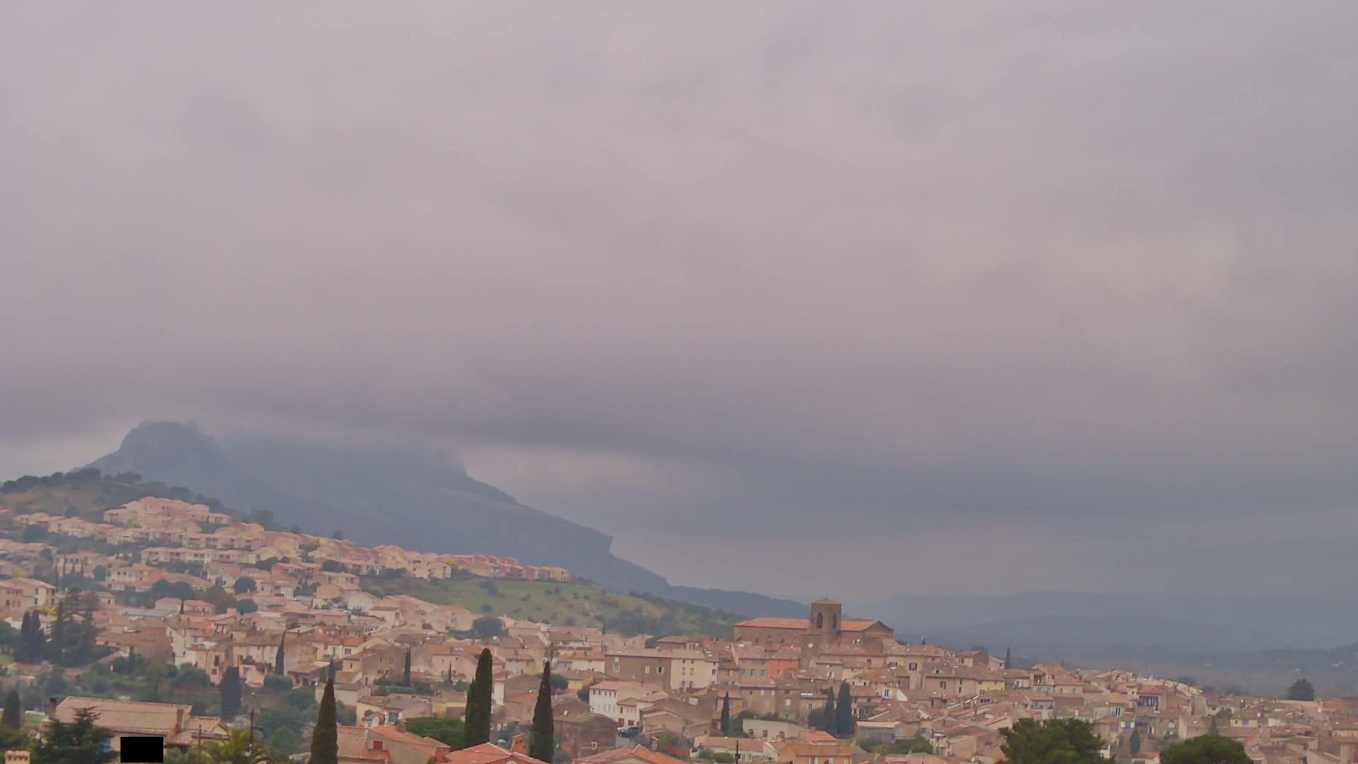 A sprawling town of tan-colored buildings sits on a hillside beneath an overcast sky, with a mountain range visible in the background.