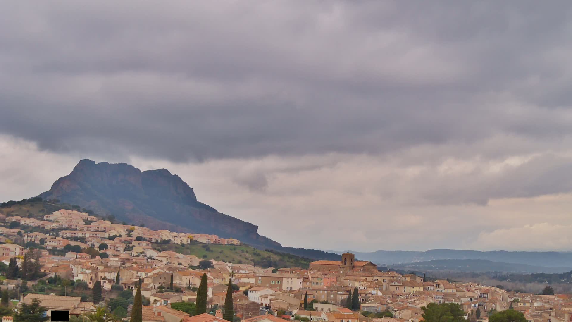 A hillside town of tan and brown buildings nestles beneath a large, dark mountain under a cloudy sky.