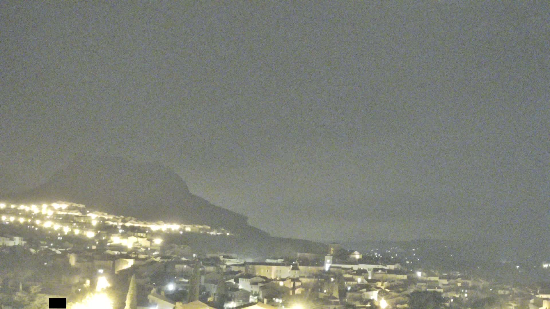 A nighttime view of a town nestled against a hazy mountain under a muted sky.