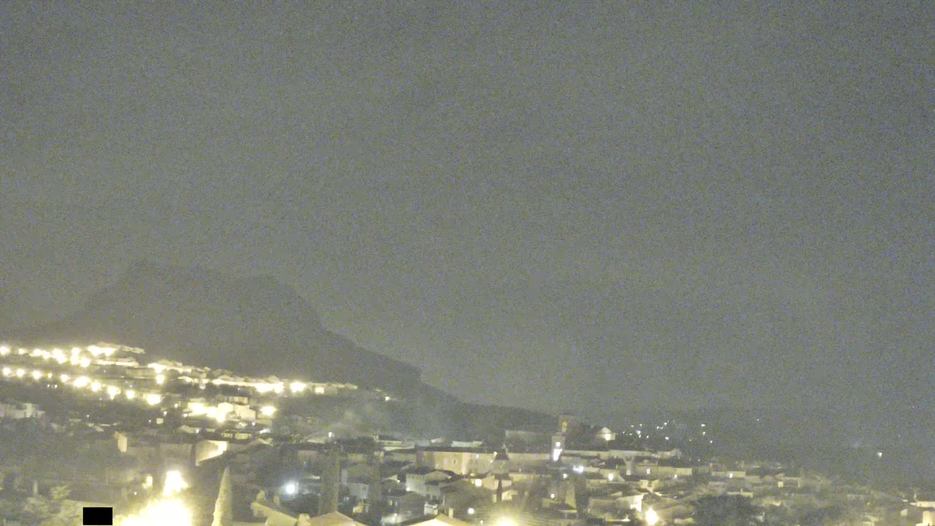 A nighttime view of a town nestled against a hill under a hazy sky.