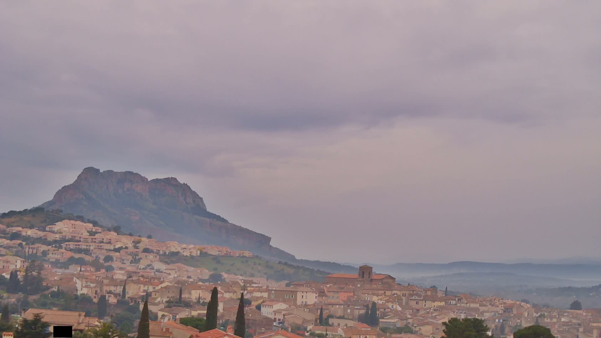 A hazy, overcast sky hangs over a town nestled at the foot of a large, dark mountain.