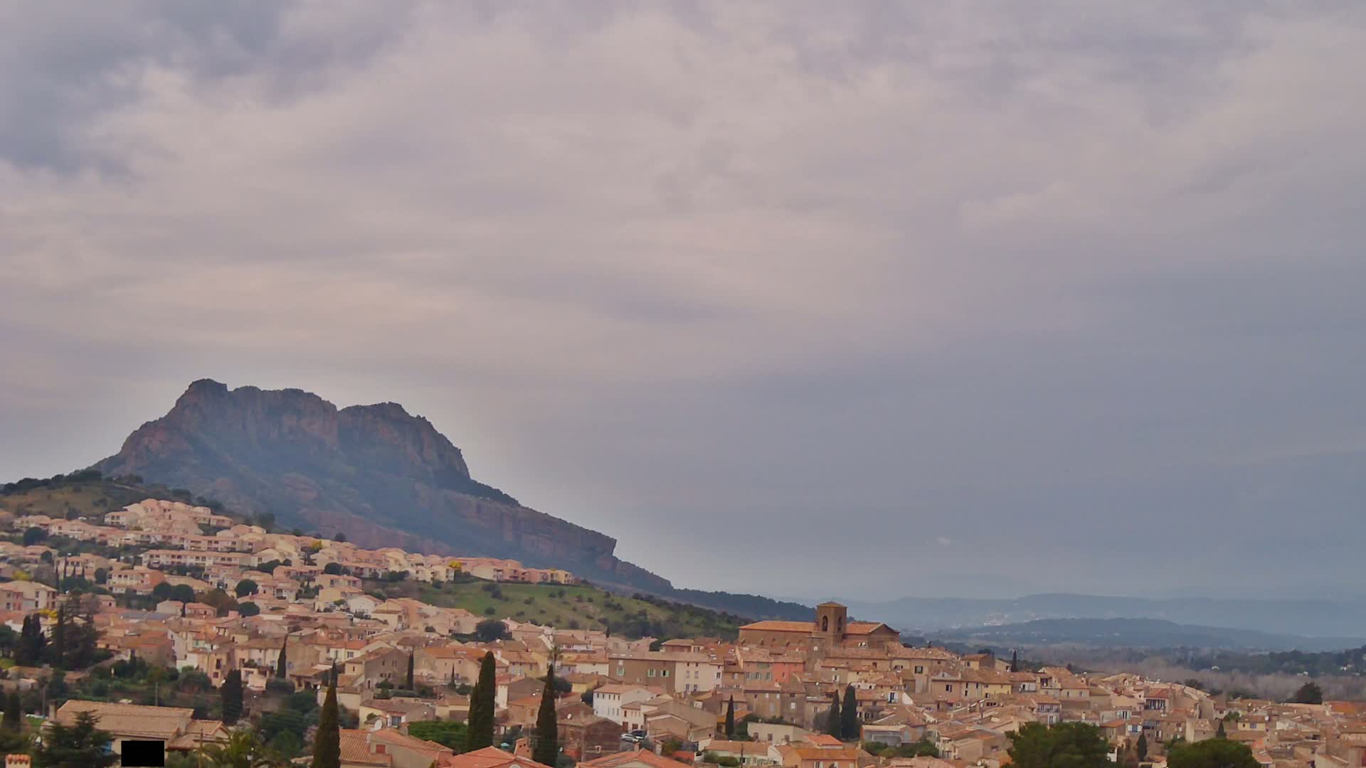 A hillside town of tan and brown buildings nestles against a large, dark mountain under a cloudy sky.