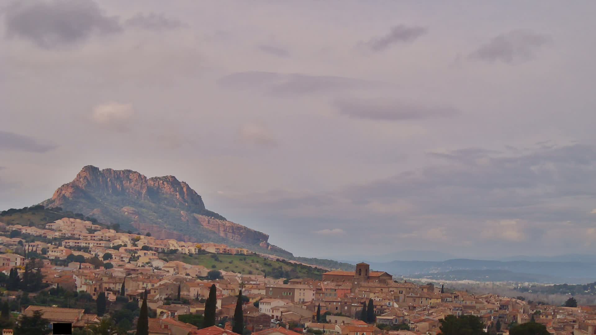 A town of tan and brown buildings sits on a hillside beneath a large, rocky mountain under a cloudy sky.