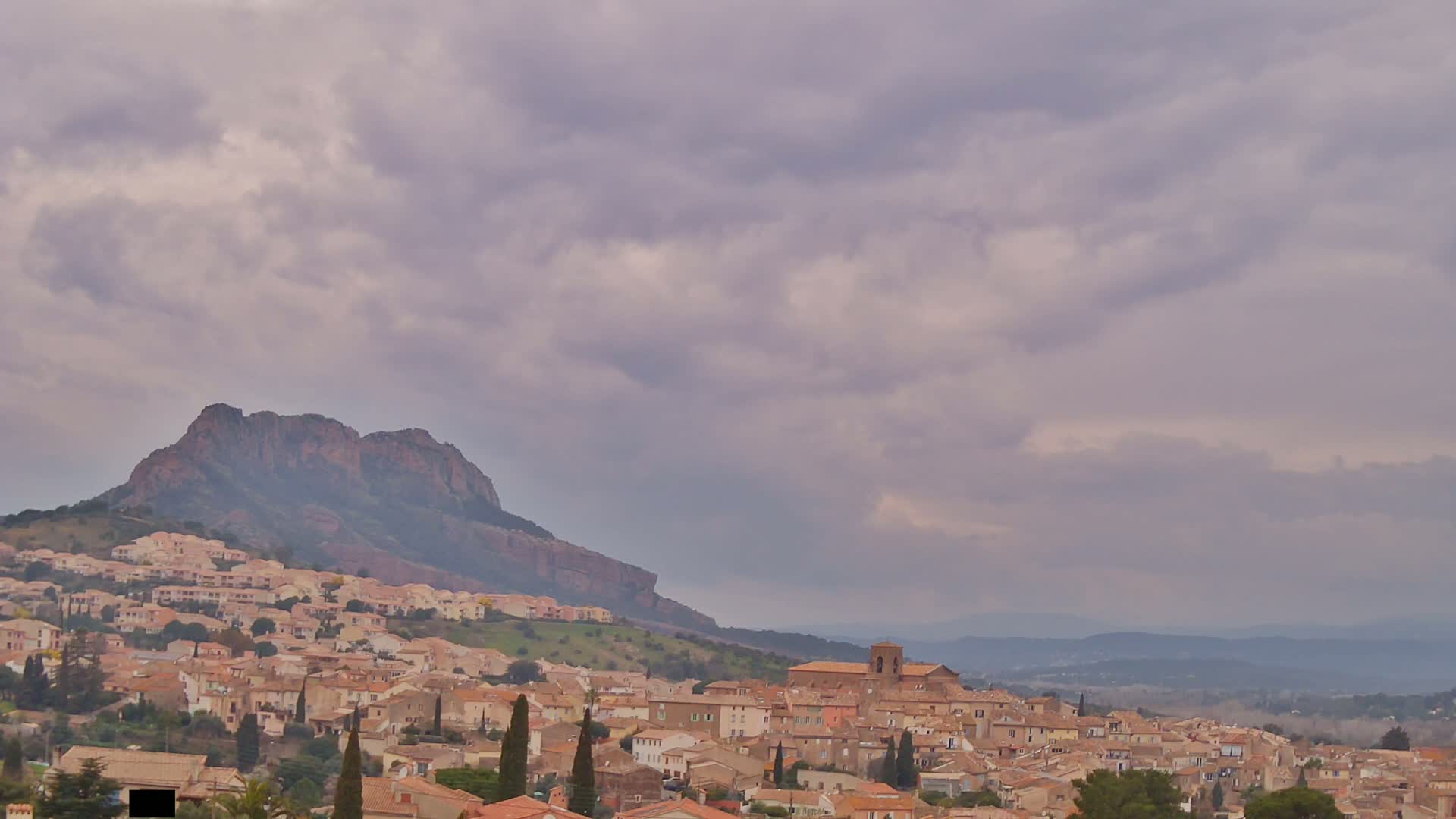 A hilltop town with tan buildings nestles against a large, dark mountain under a cloudy sky.