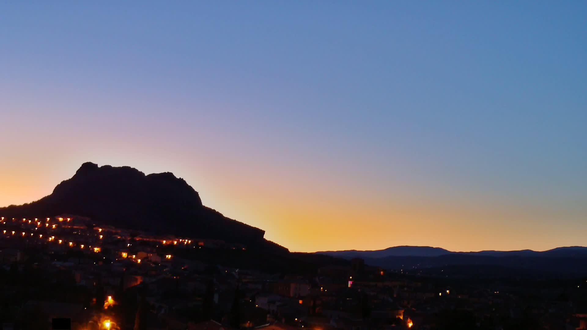 A town nestled against a large, dark mountain silhouetted against a sunset sky.