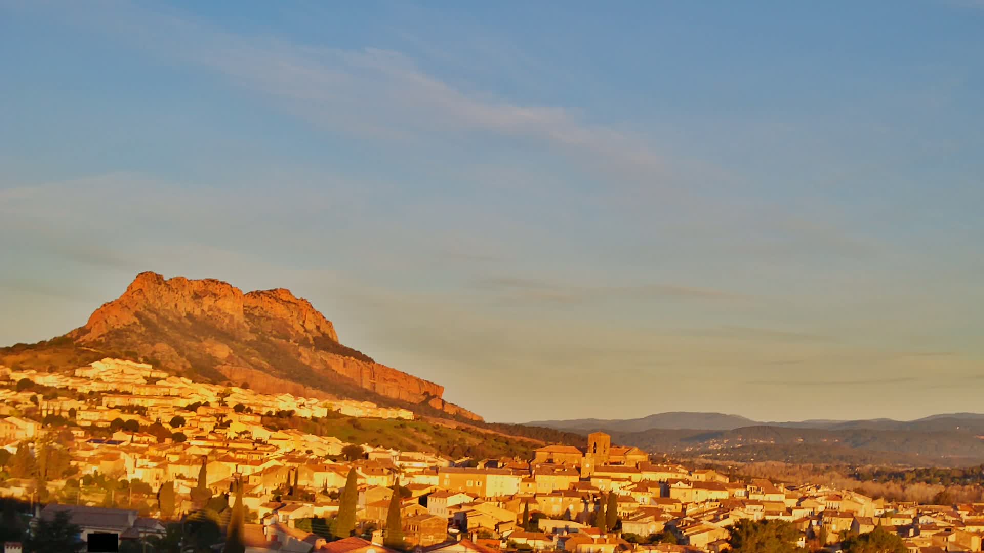 A town of tan buildings clusters at the base of a large, sunlit, reddish-brown cliff, with additional hills visible in the background under a mostly clear, pale-yellow sky.