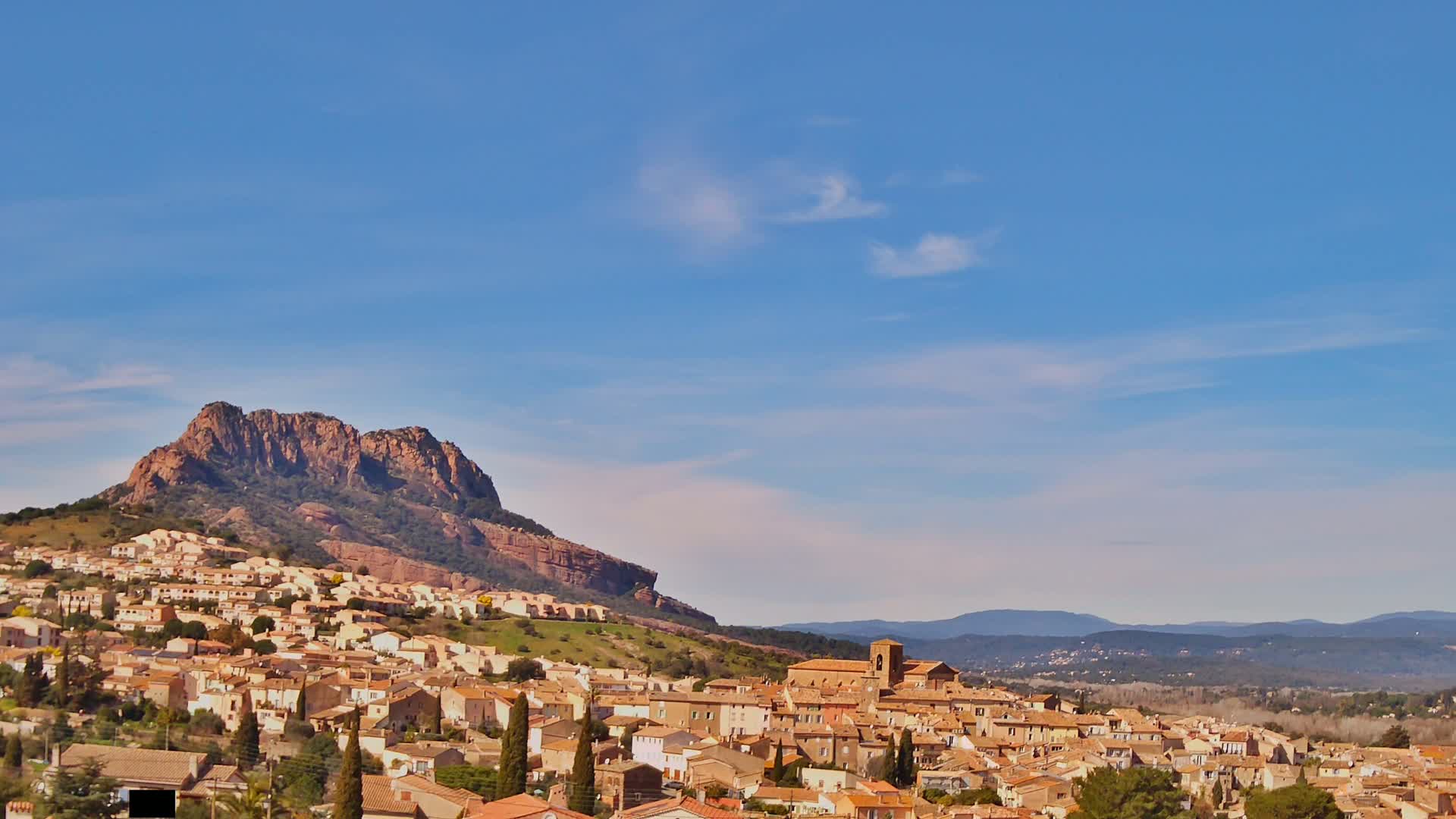 A tan-colored town with a church is nestled at the base of a large, rocky mountain under a clear blue sky.