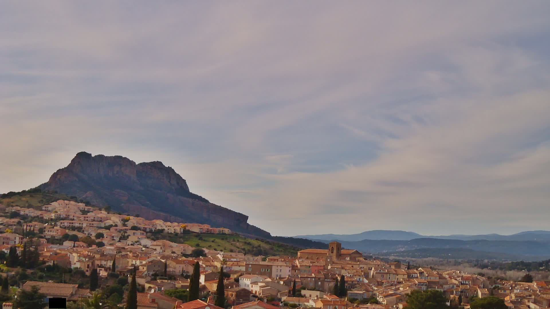 A large hill rises behind a sprawling town of tan and brown buildings under a mostly cloudy sky.