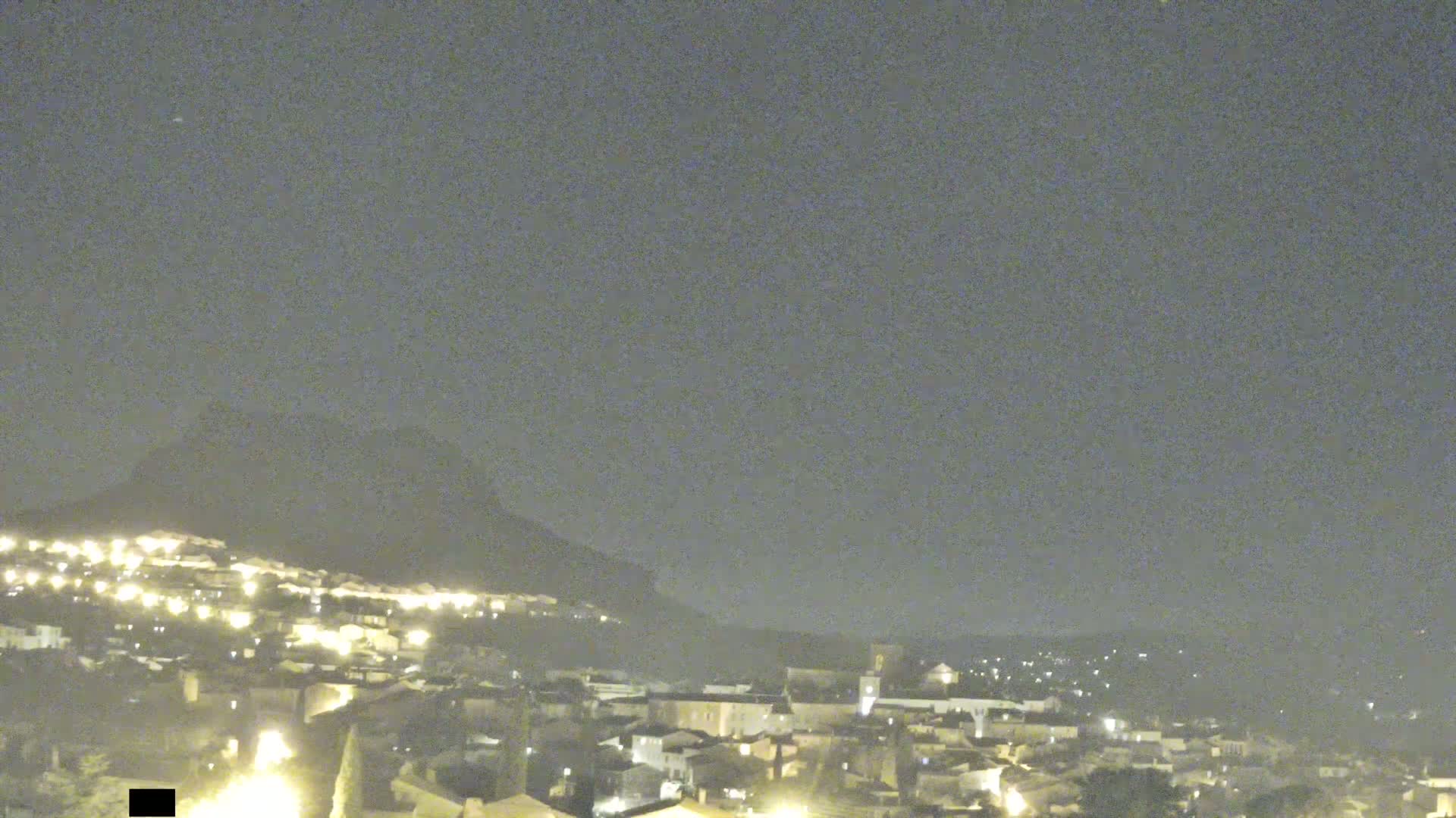 A nighttime long shot shows a hilltop town illuminated by lights against a hazy, dark sky.