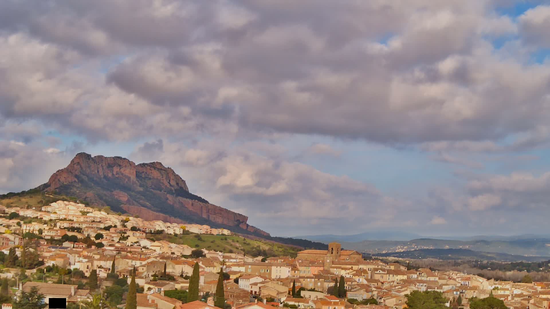 A sprawling town of tan and brown buildings nestled against a large, reddish-brown mountain under a partly cloudy sky.