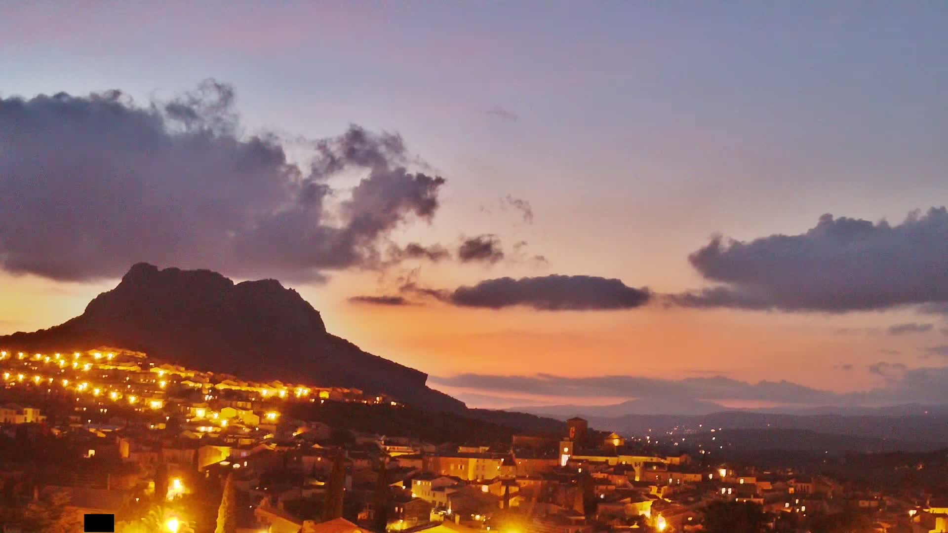A hilltop town illuminated at dusk, nestled against a silhouetted mountain under a partly cloudy, orange and purple sky.