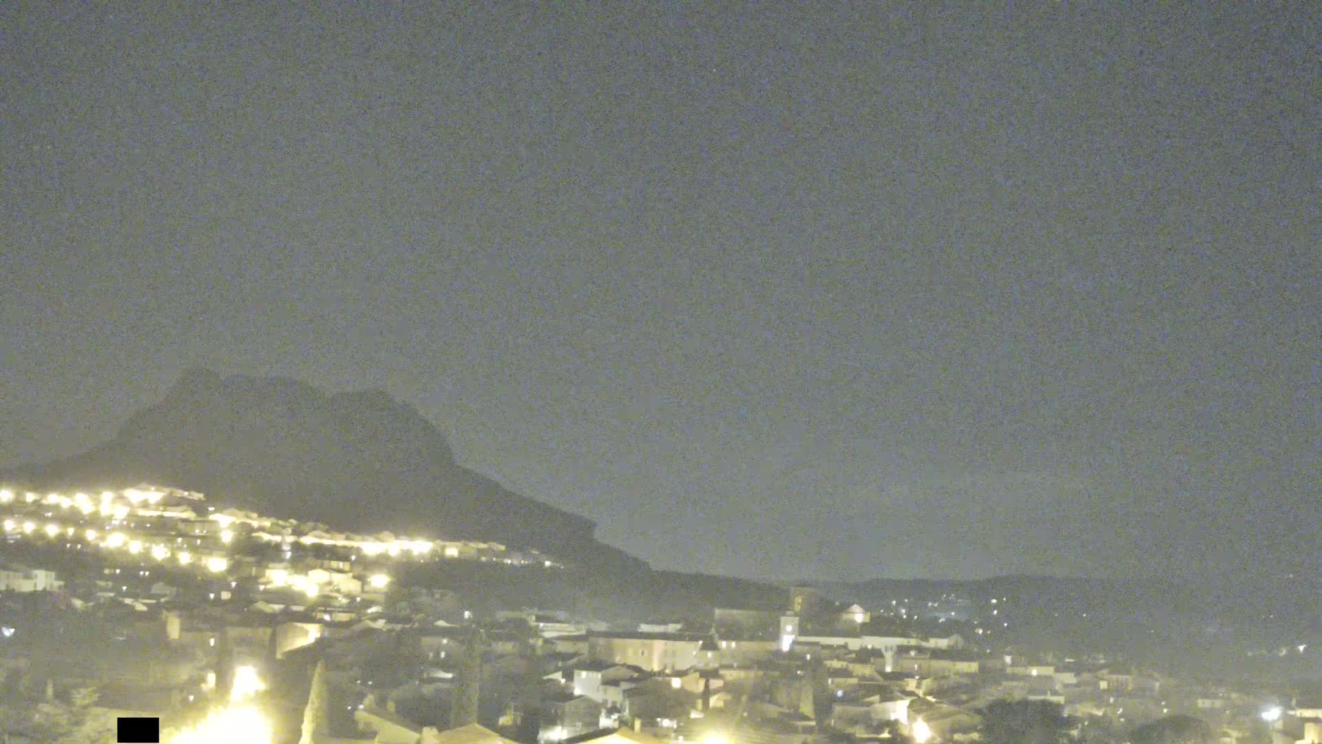 A nighttime view of a town illuminated by lights, nestled against a dark mountain under a cloudy sky.