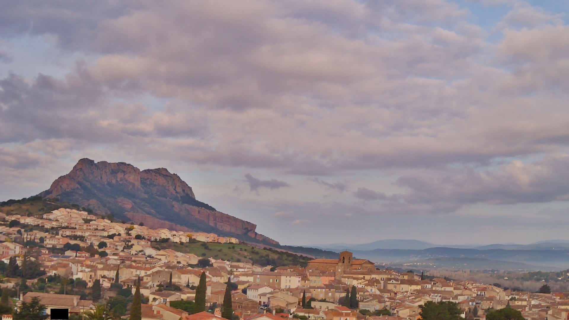 A large hill, topped with a rocky mountain, overlooks a town of low, tan buildings under a mostly cloudy sky.
