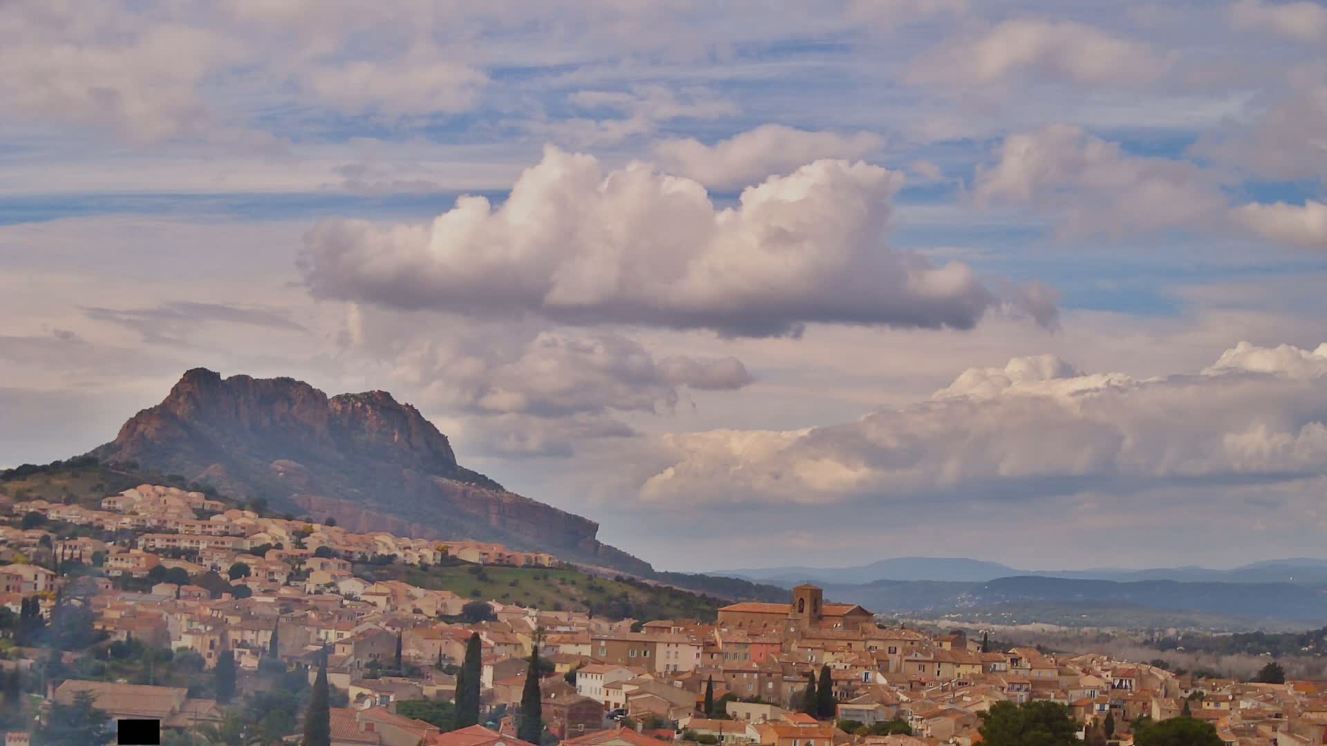 A hilly town with tan buildings nestles beneath a large, dark-colored mountain under a partly cloudy sky.