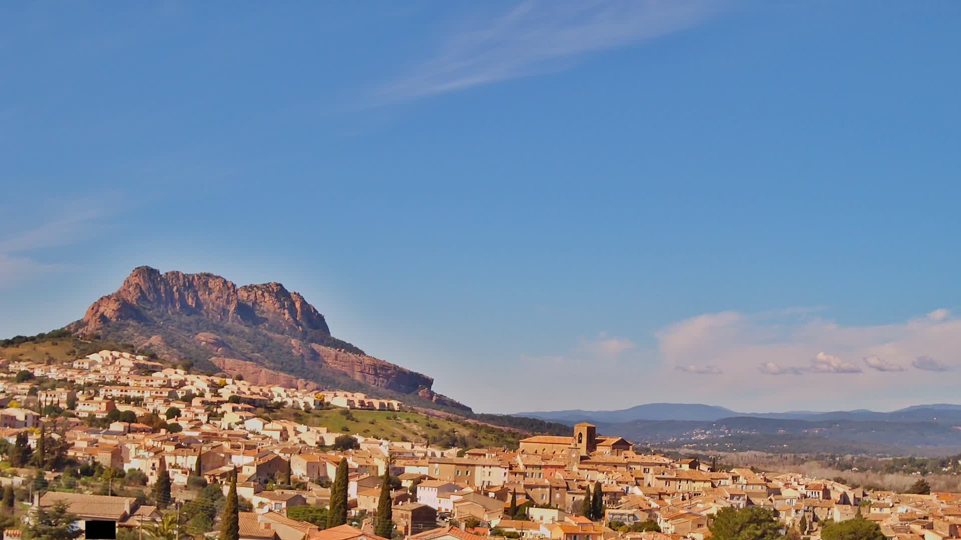 A village of tan-colored buildings sprawls across a hillside beneath a large, rocky mountain under a clear blue sky with a few wispy clouds.