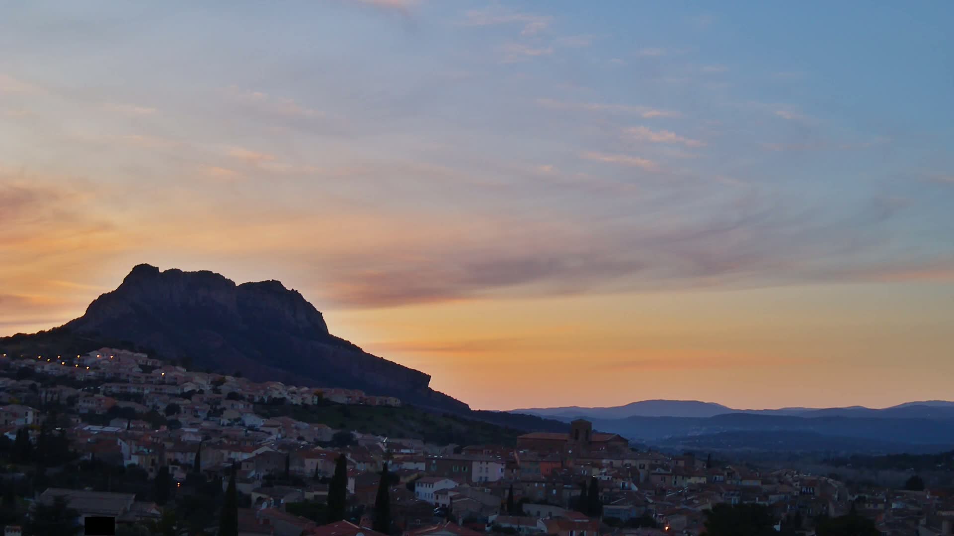 A village nestled at the foot of a large, dark mountain under a pastel sunset sky.