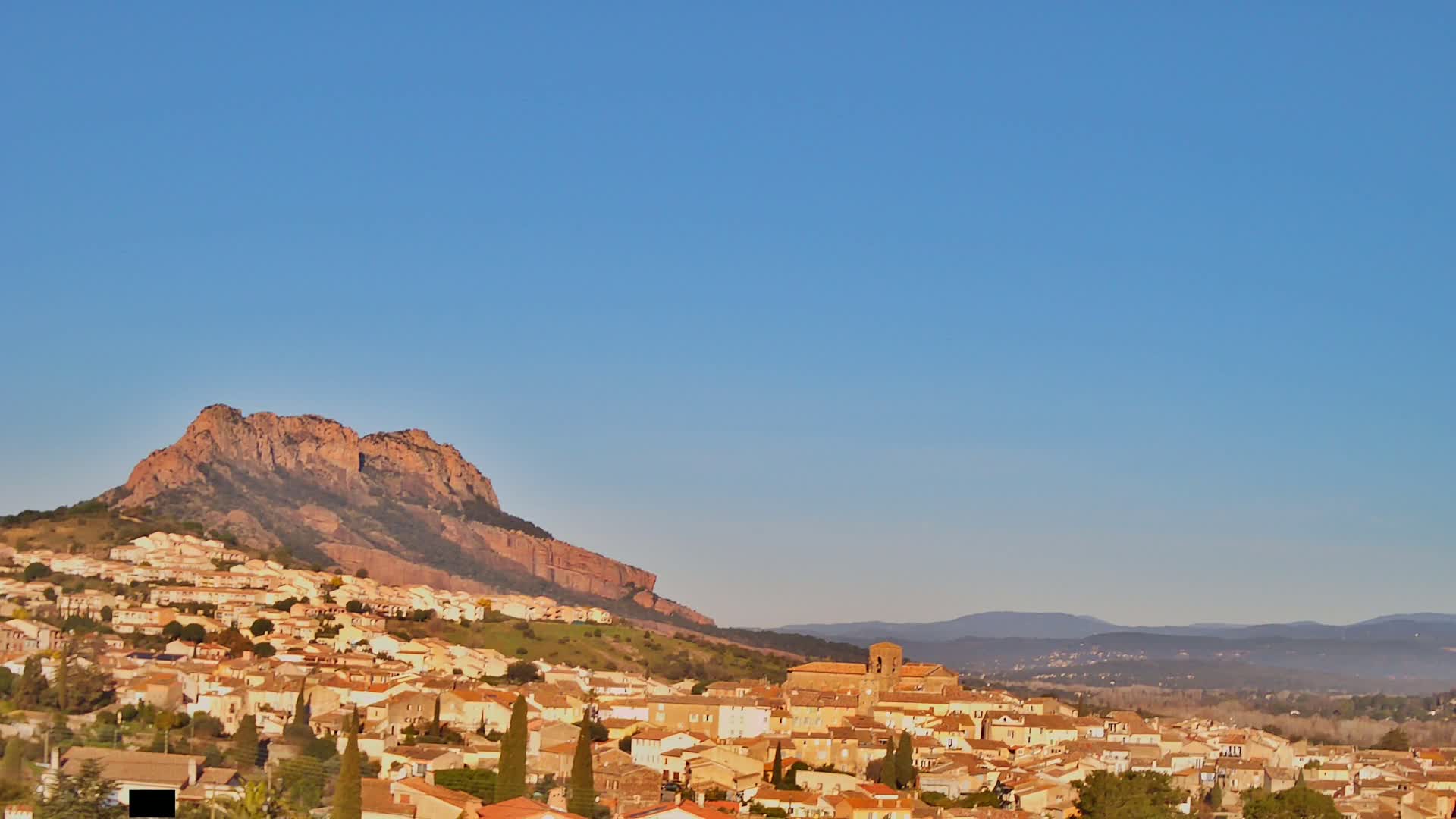 A village of tan-colored buildings nestled at the base of a large, reddish-brown mountain under a clear blue sky.