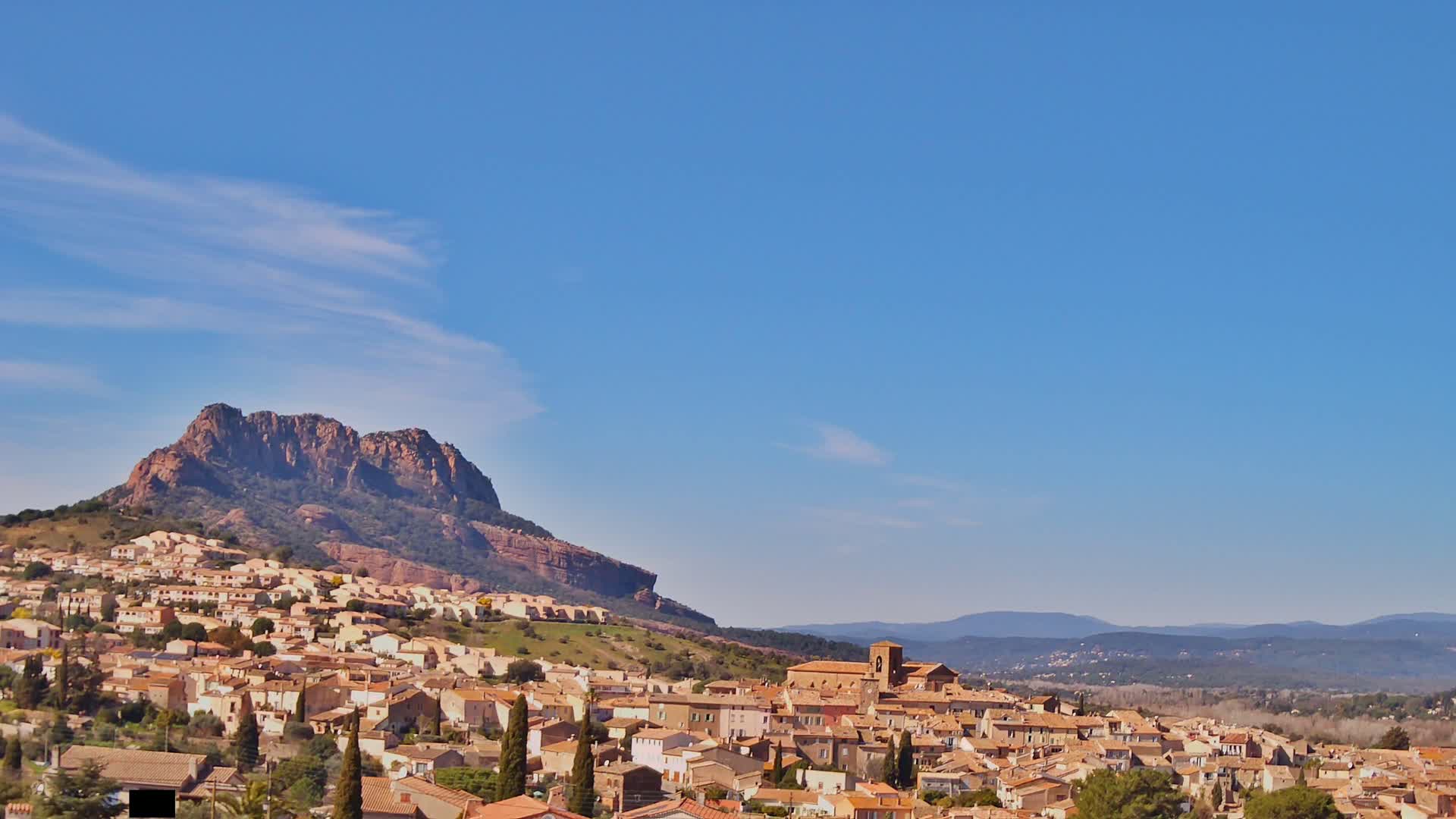A town of tan-colored buildings sprawls across a hillside beneath a large, reddish-brown mountain against a clear blue sky.