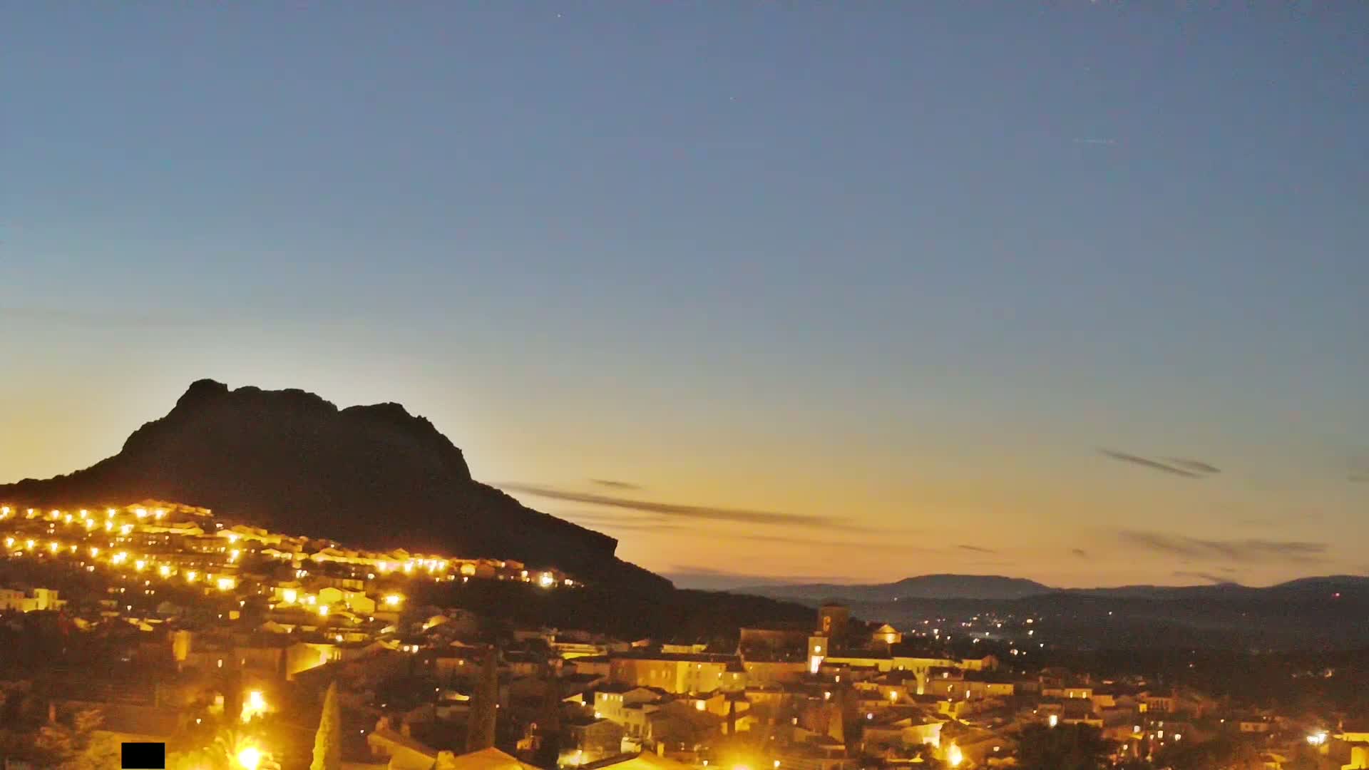 A nighttime view of a town illuminated by lights, nestled against a large, dark mountain under a twilight sky.