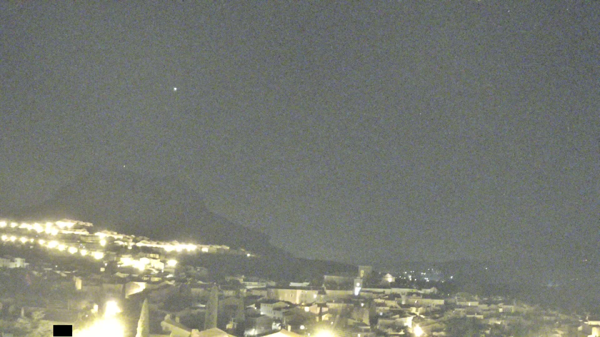 A nighttime long shot shows a hilltop town illuminated with lights under a dark, clear sky.