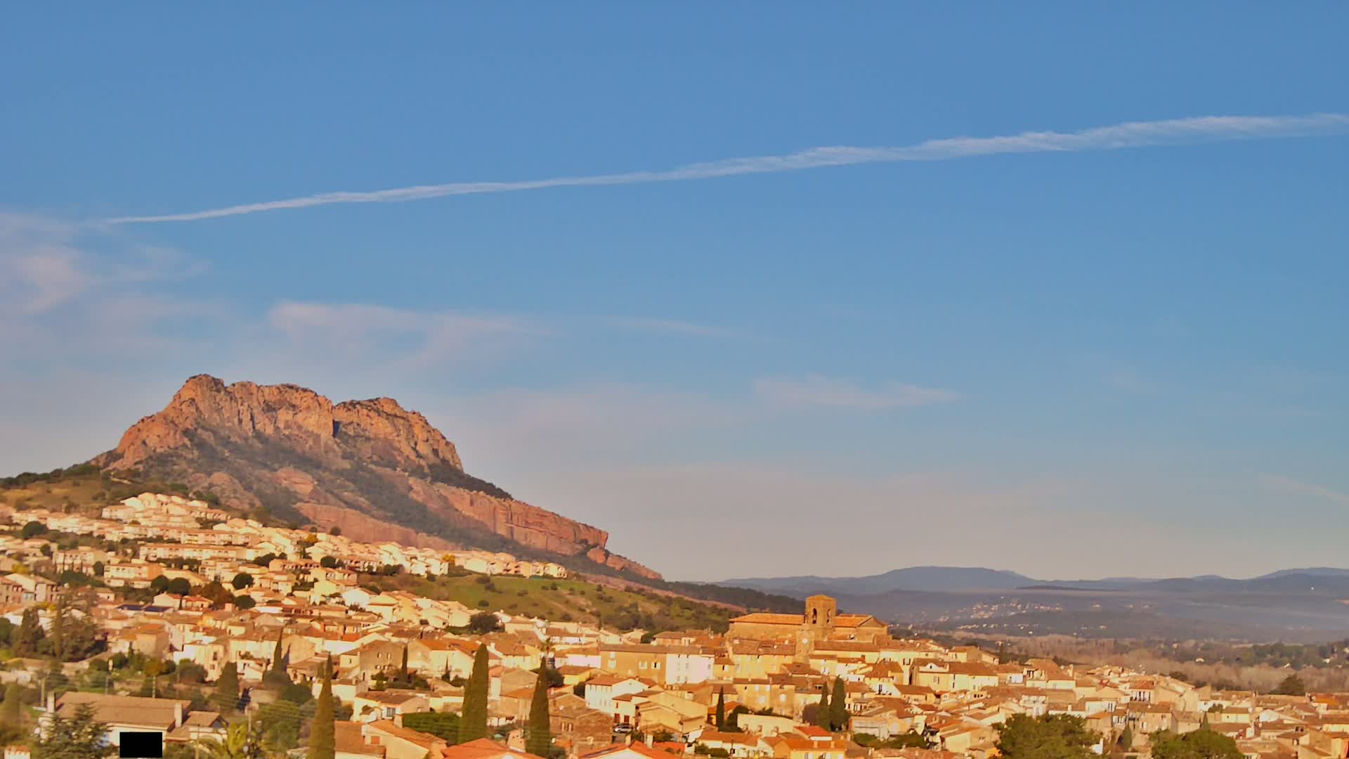 A town of tan-colored buildings nestled against a large, reddish-brown mountain under a clear, blue sky with a faint contrail.