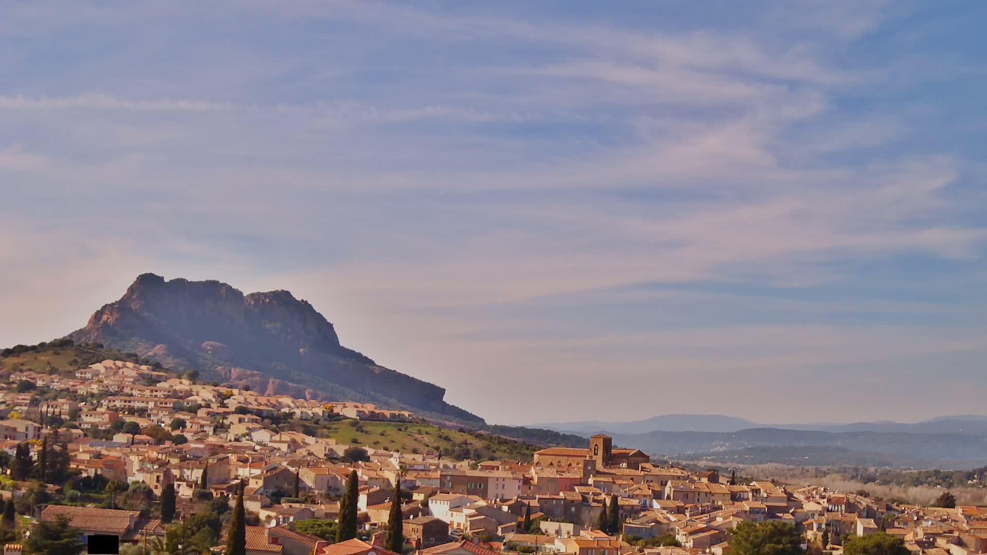 A small town of tan-colored buildings nestled against a large, dark mountain under a mostly clear, light blue sky.