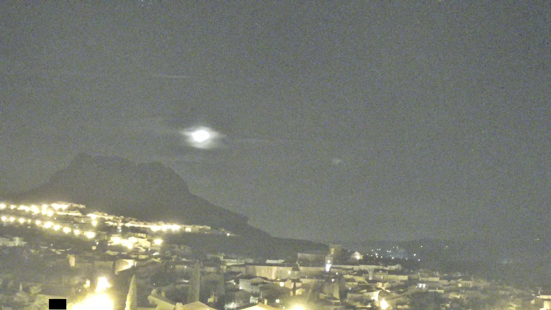 A nighttime view of a town nestled at the base of a hill, illuminated by lights and a bright moon in a mostly clear sky.