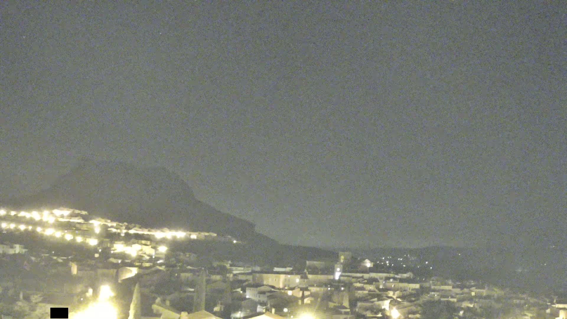 A nighttime, long shot of a town nestled at the base of a hill under a dark, clear sky.