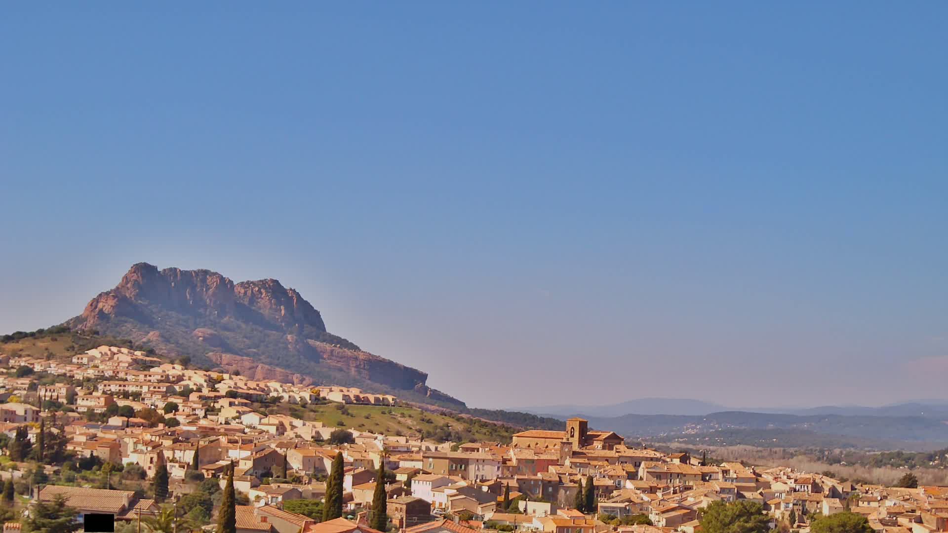A town of tan and light brown buildings clusters at the base of a large, reddish-brown mountain under a clear blue sky.