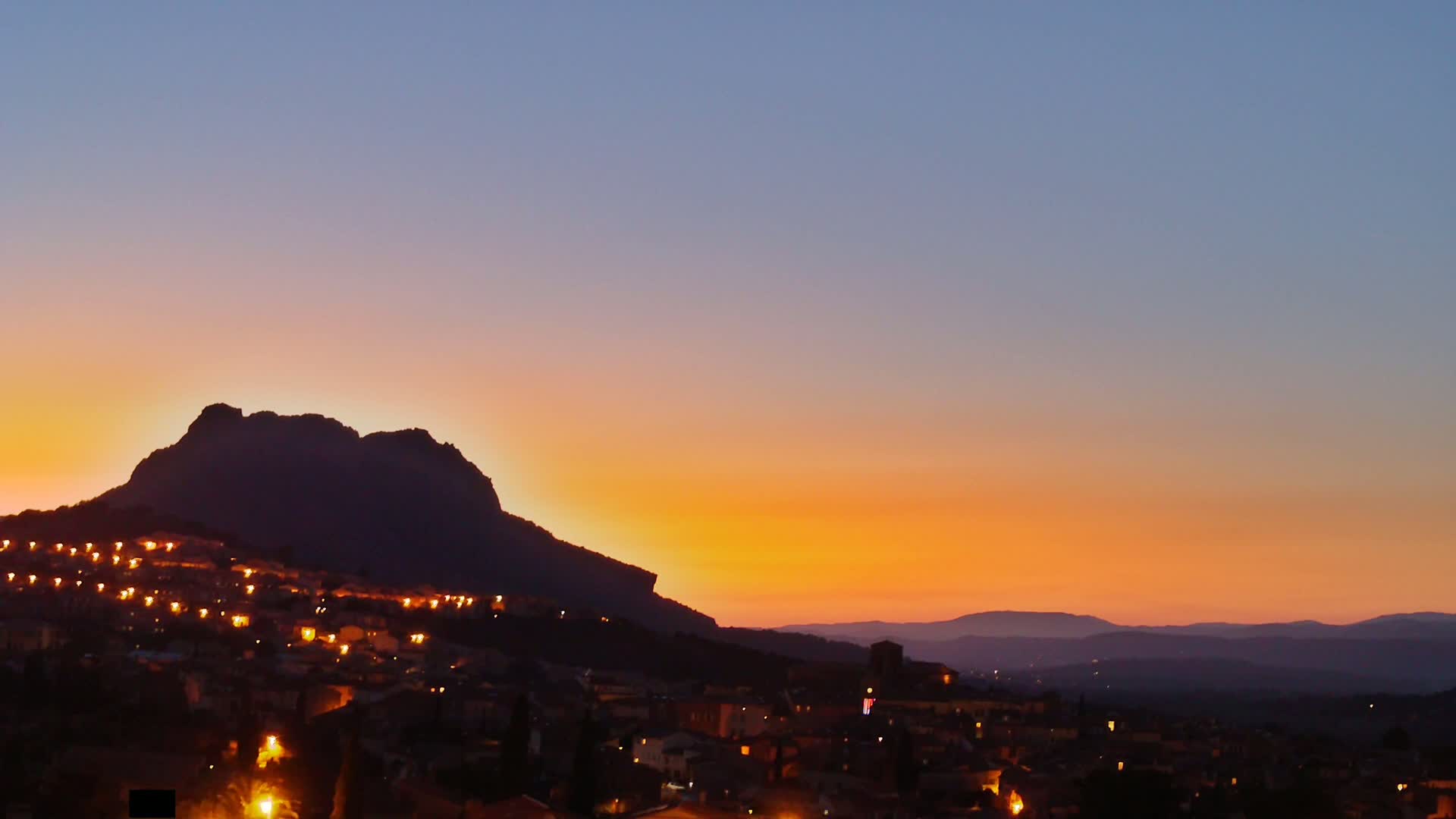 A town nestled against a silhouetted mountain at sunset, under a clear, orange and purple sky.