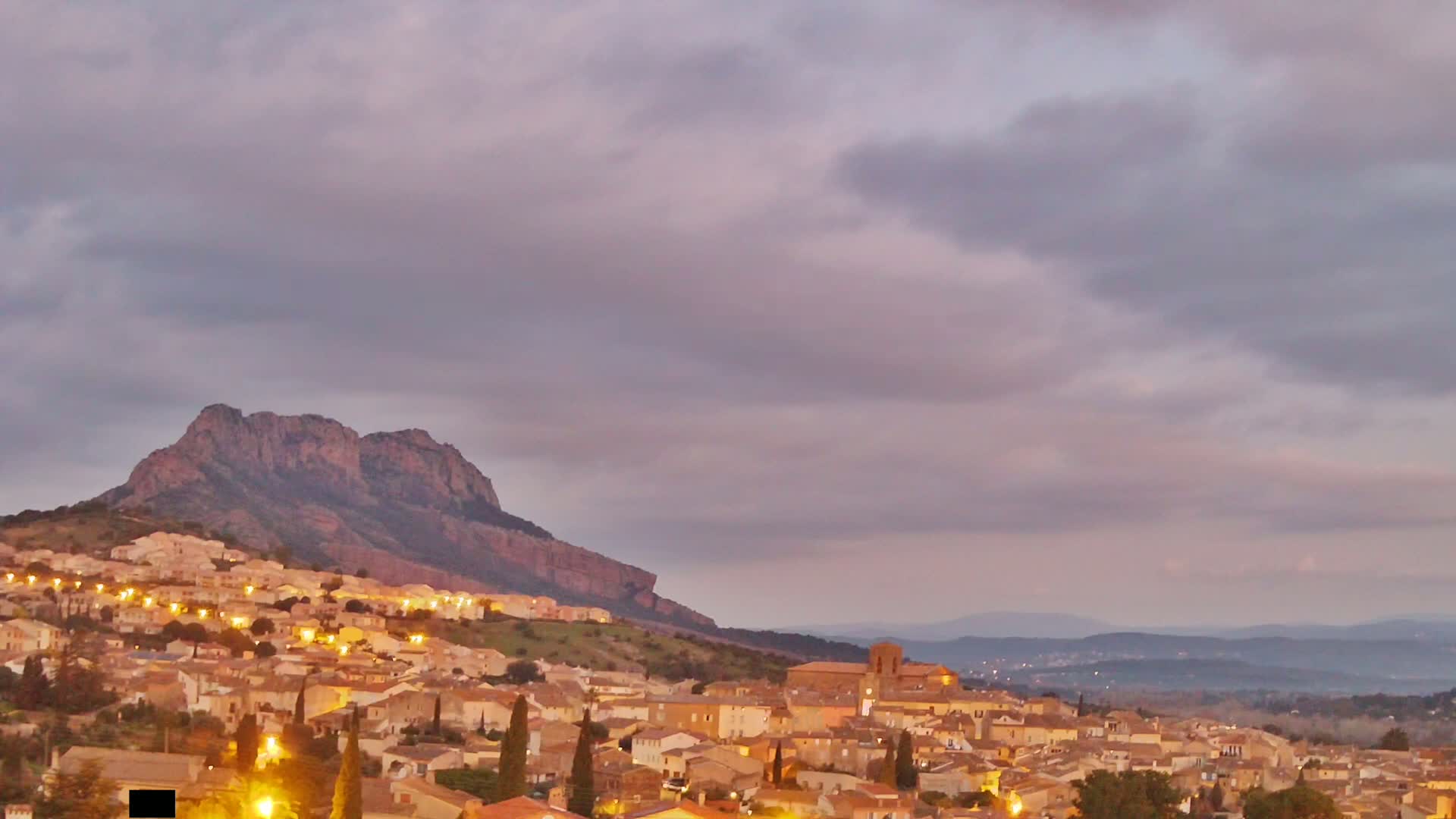 A town with illuminated buildings nestled at the base of a large, rocky mountain under a cloudy, twilight sky.
